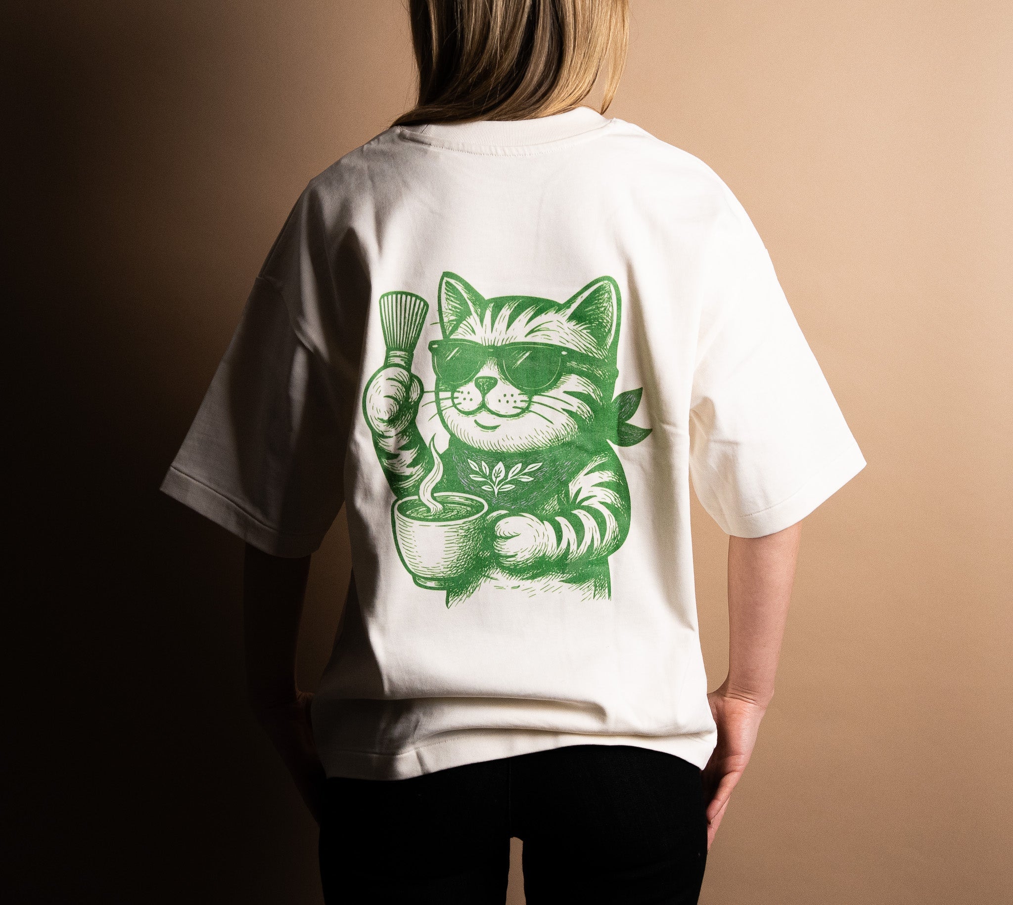 Person wearing white t-shirt with green matcha cat cartoon holding a whisk and tea bowl