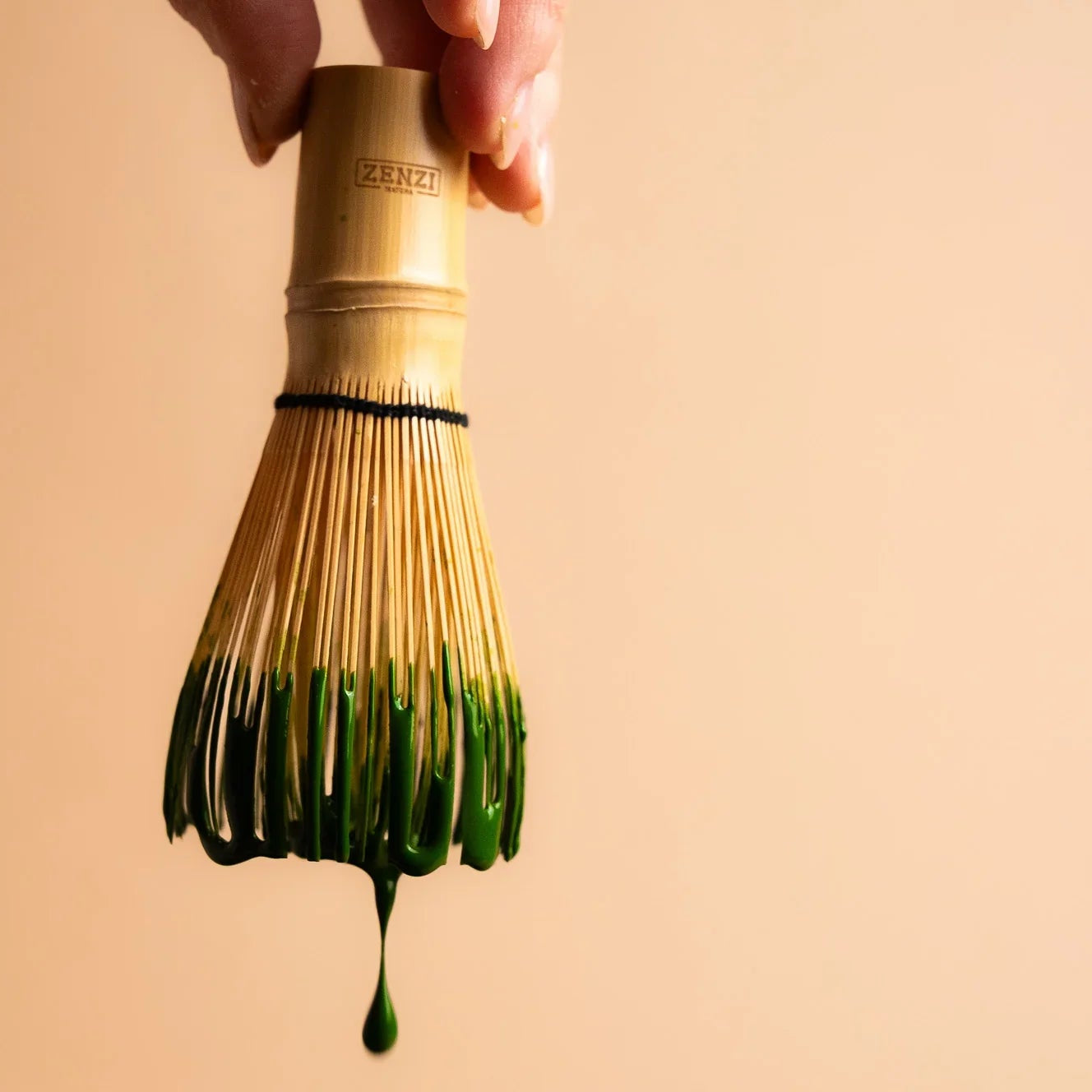 Bamboo matcha whisk with green matcha tea dripping, held by hand on beige background